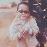Vintage photo of Aaron as a child wearing sunglasses and a fur coat, smiling at the camera.